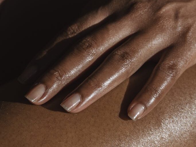 Crop anonymous African American female touching bare leg with hand while sitting on black background in room with bright sunlight